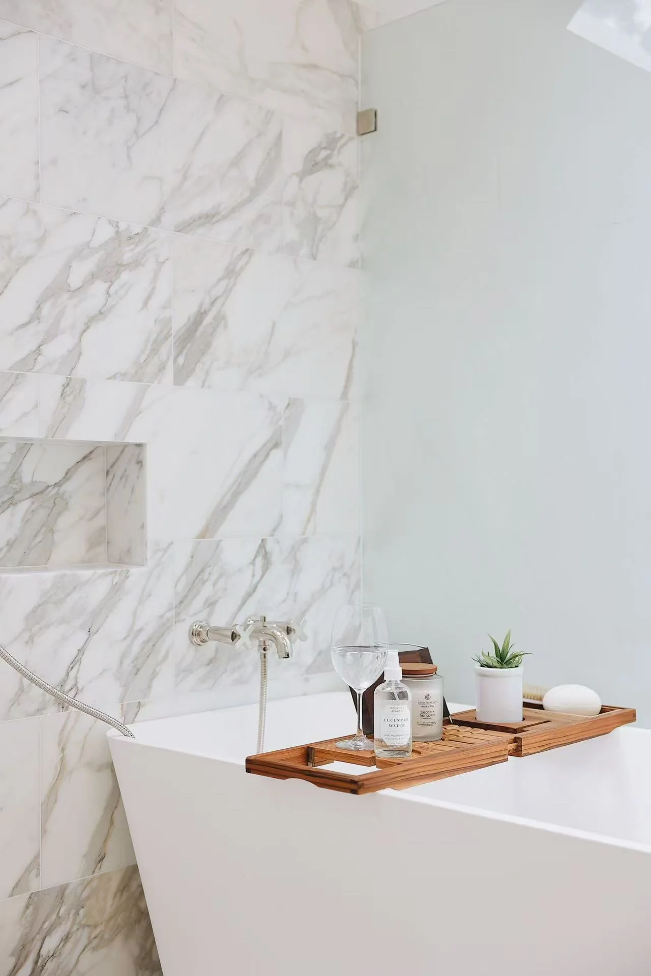 Freestanding bathtub with marble-effect wall, glass panel, and warm wood accessory